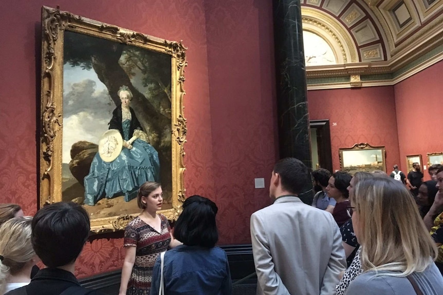 Young woman standing in front of 19th c painting, talking - with semi-circle of people around her, listening.