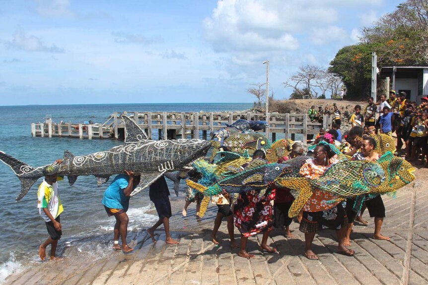 Lots of Torres Strait Islanders in front of a wharf carrying large sea creatures made of ghost nets