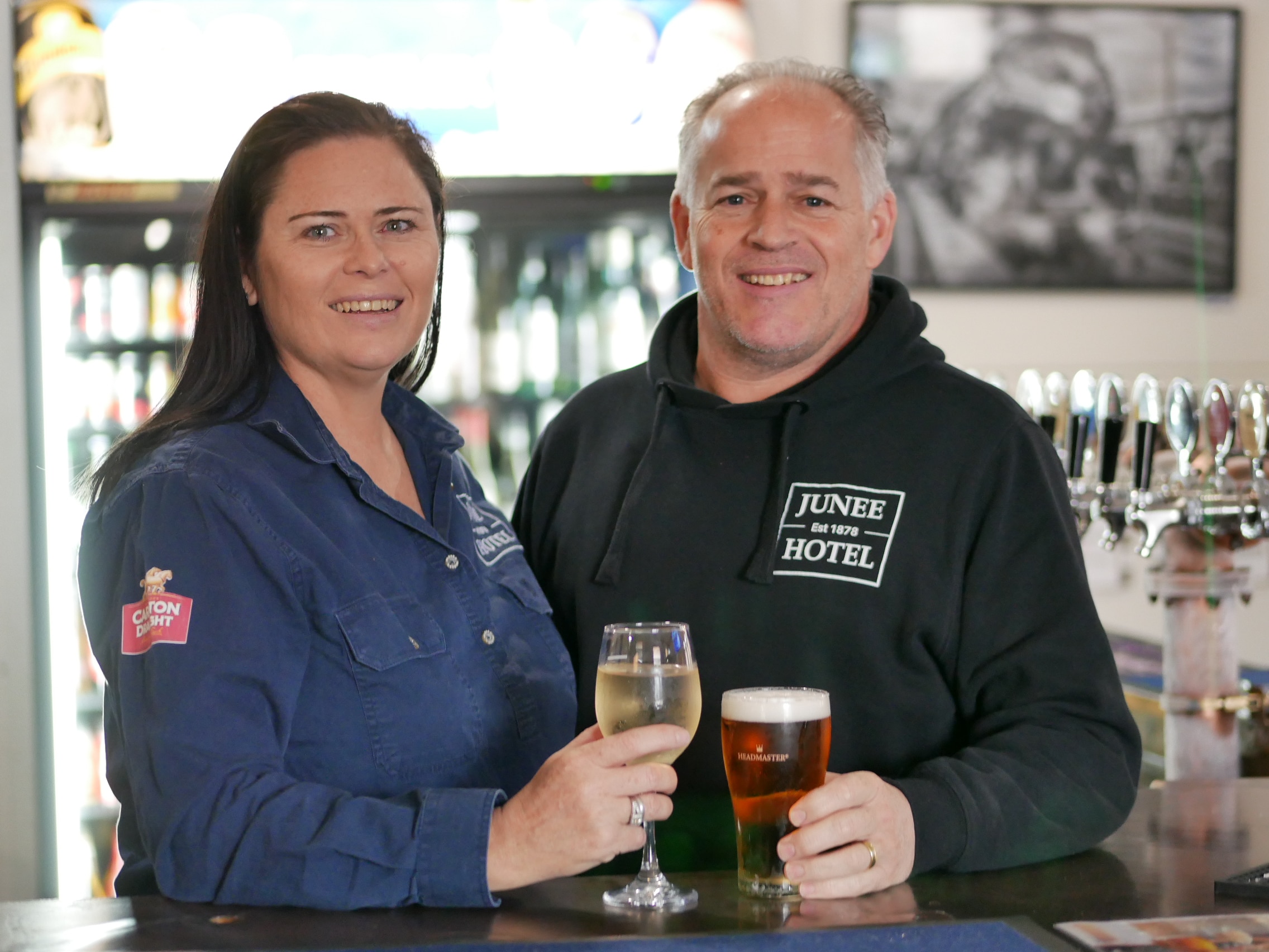 A woman holding a wine and man holding a beer standing at a bar smiling.