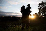 Silhouette of a woman holding her baby outside as the sun sets.