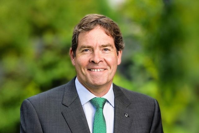 Oliver Yates, wearing suit and green tie, stands in front of green tree background.