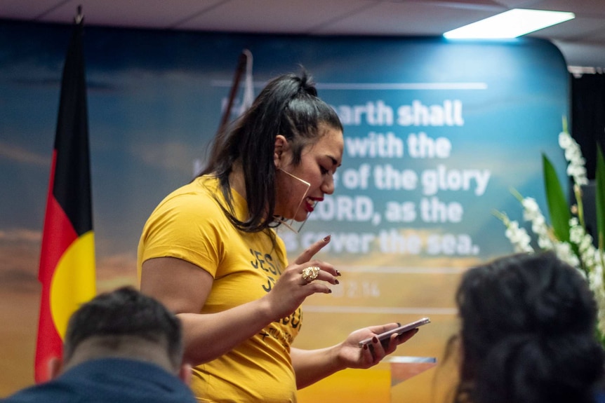 Ana Makahununiu standing in front of a small audience, is reading a sermon from her iphone.