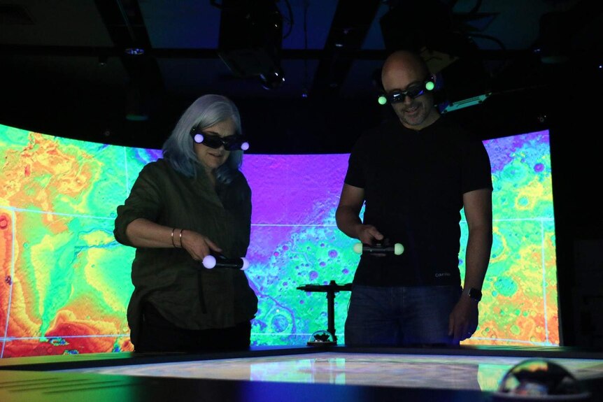 Two people wearing dark glasses, standing over a table, a colourful map of Mars in the background.