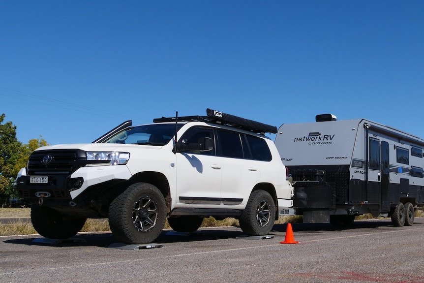 A white four wheel drive sits stationary at a truck stop with a caravan behind it and blue sky above.