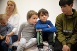 Four children sit and stare blankly. The youngest one is sitting on a woman's knee.