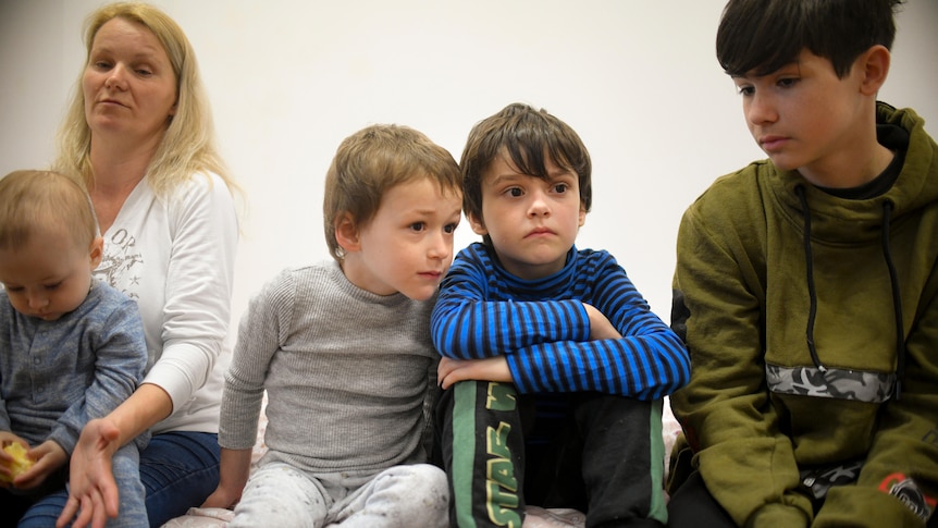 Four children sit and stare blankly. The youngest one is sitting on a woman's knee.