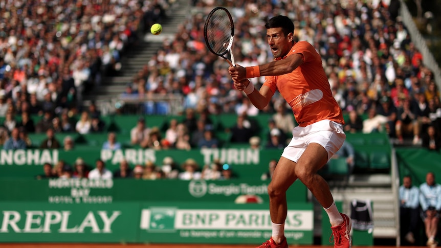 Novak Djokovic stands on the red clay and braces himself, mouth open, as he hits a backhand during a match.