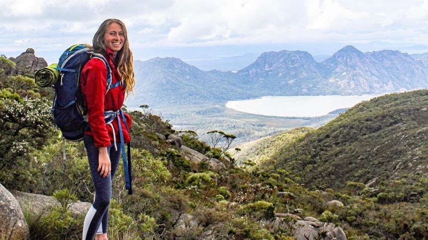 A woman with long hair smiling in a red fleece, navy leggings and a hiking pack, with a mountain range and ocean behind her.