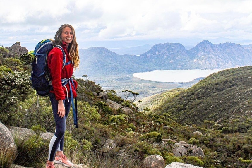A woman with long hair smiling in a red fleece, navy leggings and a hiking pack, with a mountain range and ocean behind her.