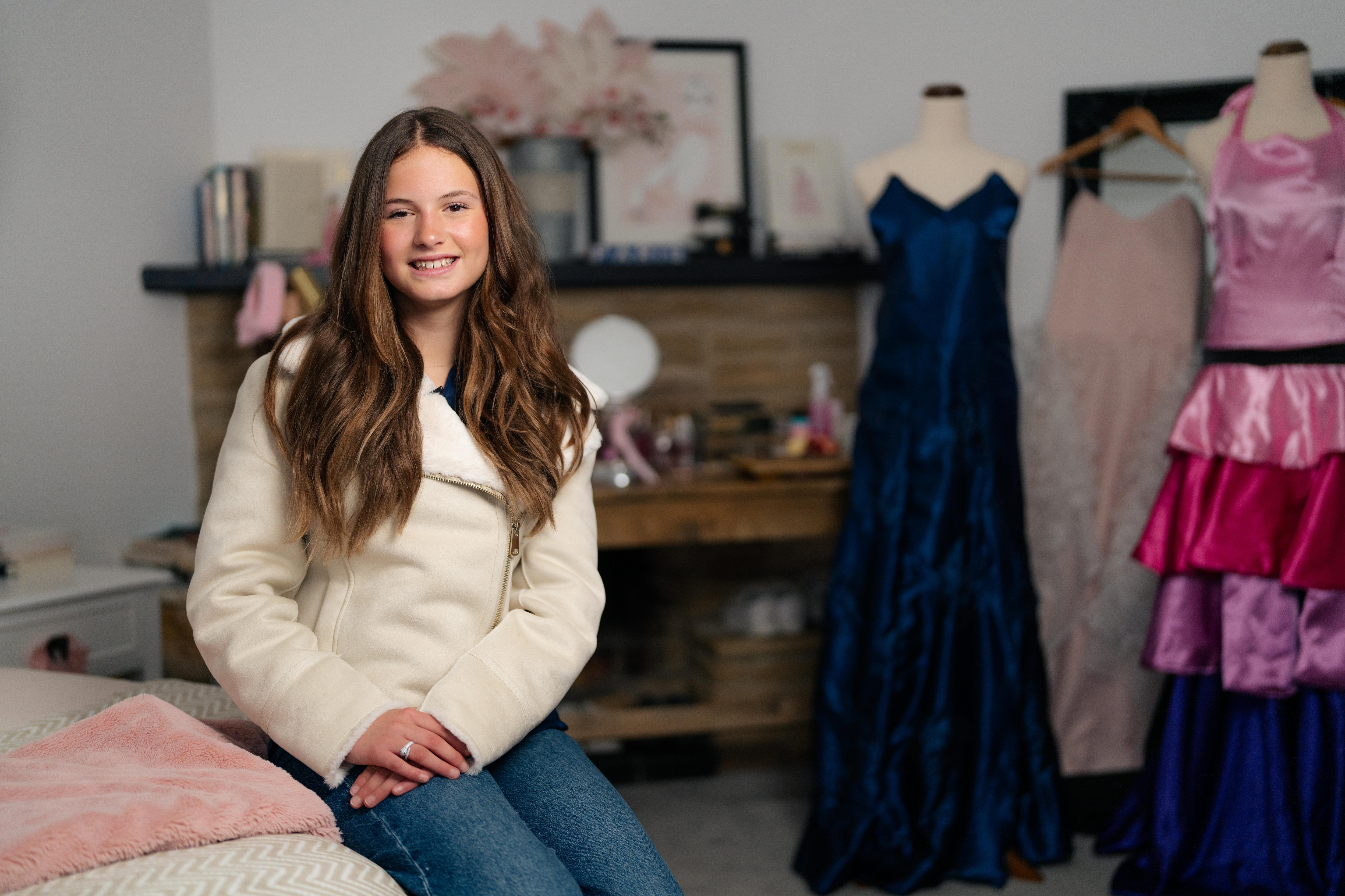 Young girl smiles for photo, sitting in her bedroom with dresses in the background
