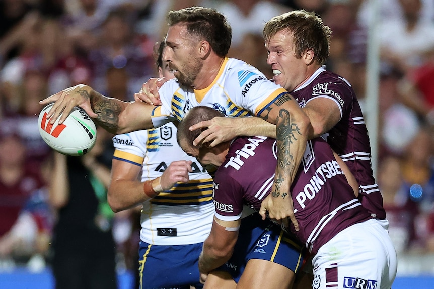 An Eels NRL player holds the ball with his right hand as he is tackled by two Manly opponents.