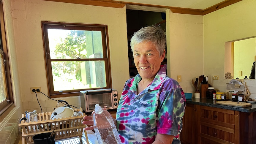 Woman with short grey hair and a colourful shirt stands at a sink holding an empty plastic dish washing liquid bottle.