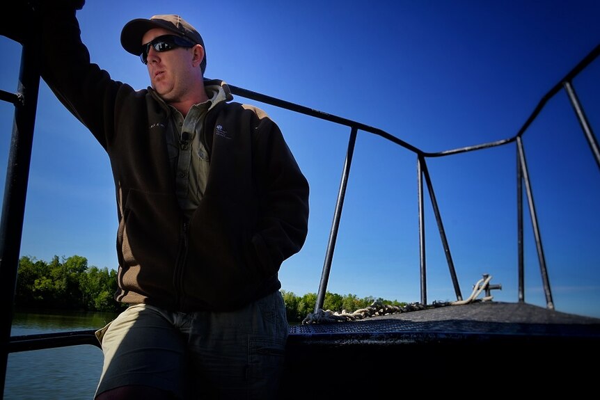 Ian Hunt wears a brown jumper, sunglasses and a brown cap, as he sits at the stern of the boat, near ocean and mangroves.
