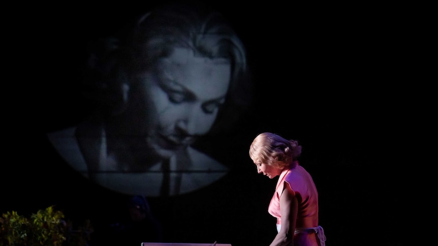 A woman in an apron prepares food in a kitchen, on stage