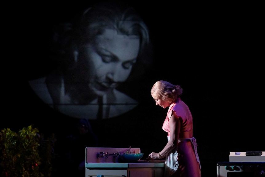 A woman in an apron prepares food in a kitchen, on stage