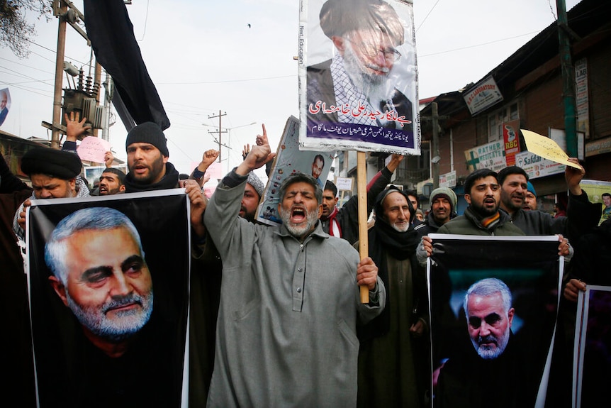 A large crowd of men carry placards showing the image of Qassem Soleimani as they raise their hands mid-shout.