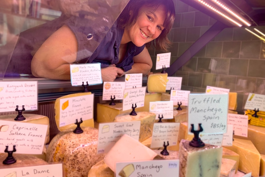 Cheese shop owner smiling leaning in through fridge window with various cheeses on display and labelled 