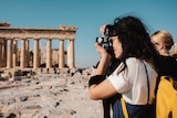 A woman with long dark hair and a yellow backpack takes a photo of the Parthenon in Athens, Greece, on a sunny day