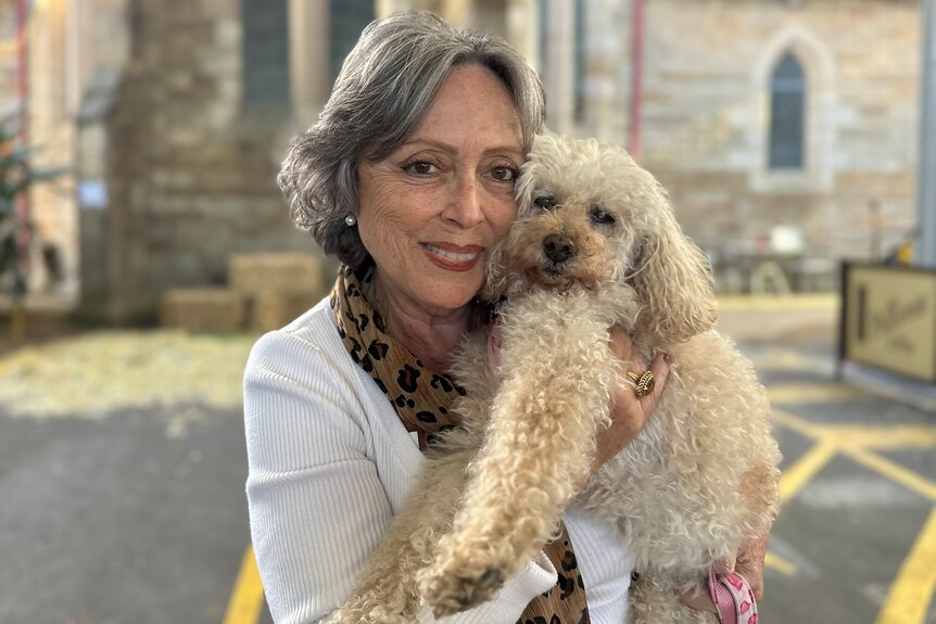 A woman holds a white scruffy dog in front of a church.