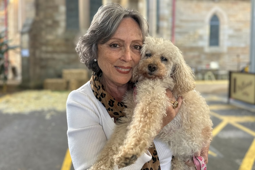 A woman holds a white scruffy dog in front of a church.