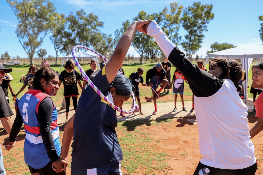 Young Aboriginal women hold hands and loop a hula hoop over their heads standing on a dusty football oval