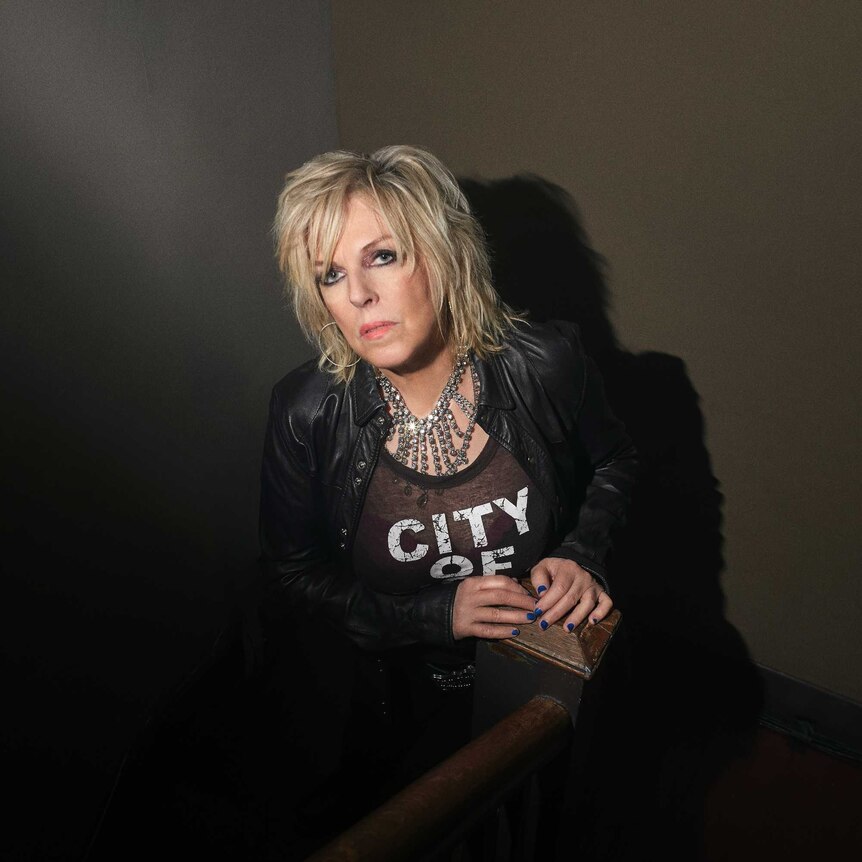 A woman in a leather jacket and bejewelled necklace sits in the corner of a stark grey room.
