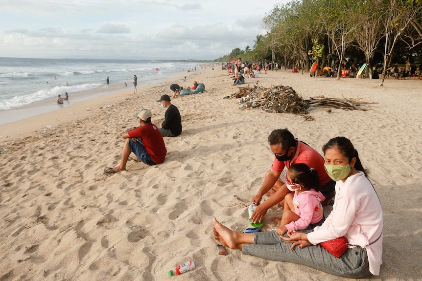 Small groups of people sit spaced apart on a beach with trees and surfboards behind them.