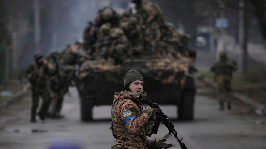 A Ukrainian solider holds a gun while looking at the camera. In the background, several soldiers are climbing on to a truck