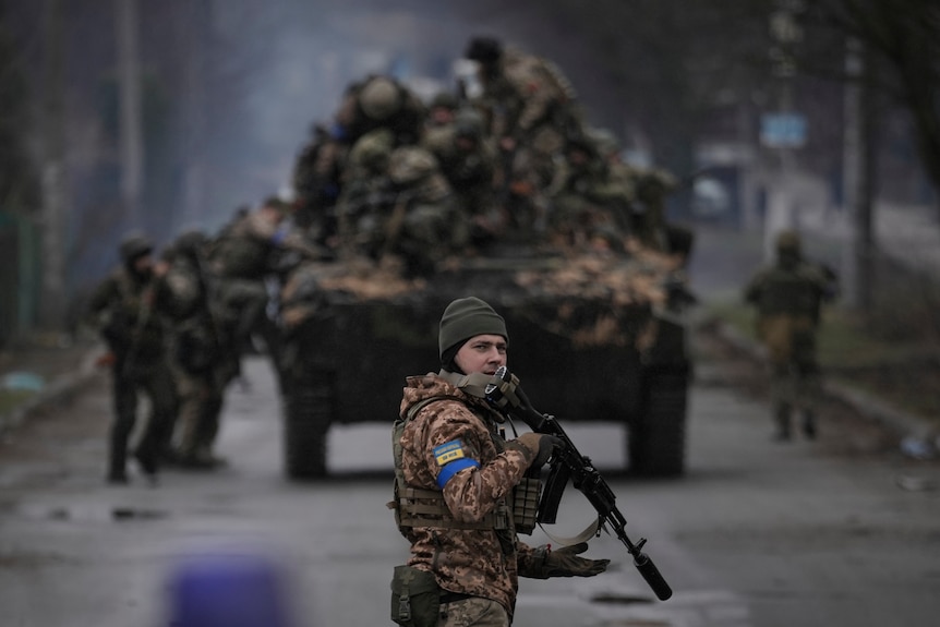A Ukrainian solider holds a gun while looking at the camera. In the background, several soldiers are climbing on to a truck