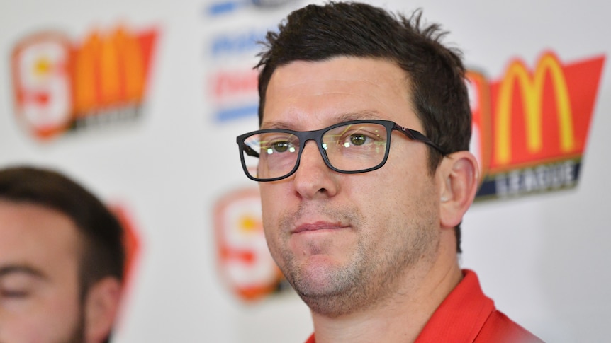 Josh Carr wears North Adelaide colours and glasses. He speaks at a press conference.