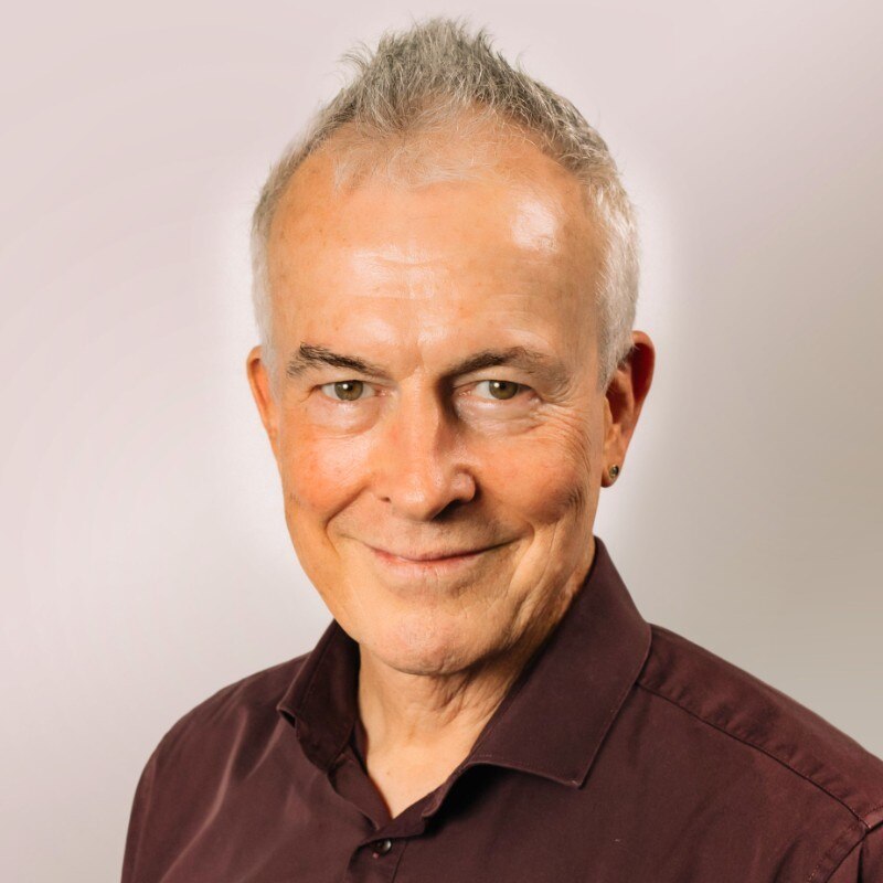 A head shot of man with grey hair smiling at the camera, with a plain background.