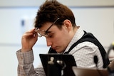 A young white man wearing bullet proof vest and sitting at table lifts his glasses as he looks down