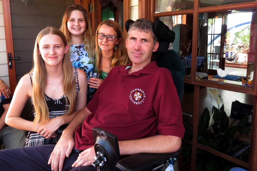 A middle-aged man in a wheelchair surrounded by his young daughters and his wife at a family home.