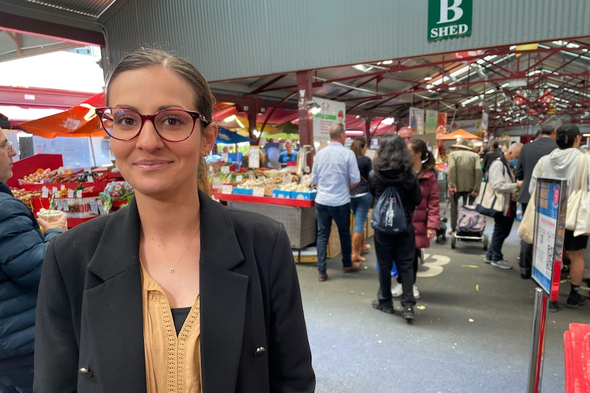 Christina Zorbas, vestida con un blazer y sonriendo a la cámara con un mercado de fondo.