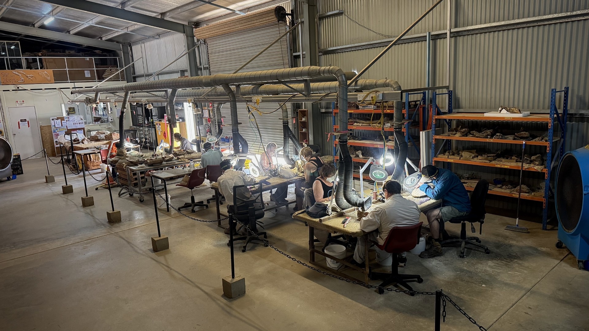 La gente se sienta en los escritorios de un gran taller con luces y ventilación colgando encima