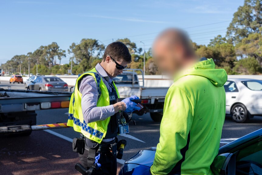 A police officers stands looking at a breathalyser screen, alongside a tow-truck driver on the side of a busy road.