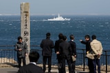 Japan Coast Guard vessel sails in the background as men watch in black suits