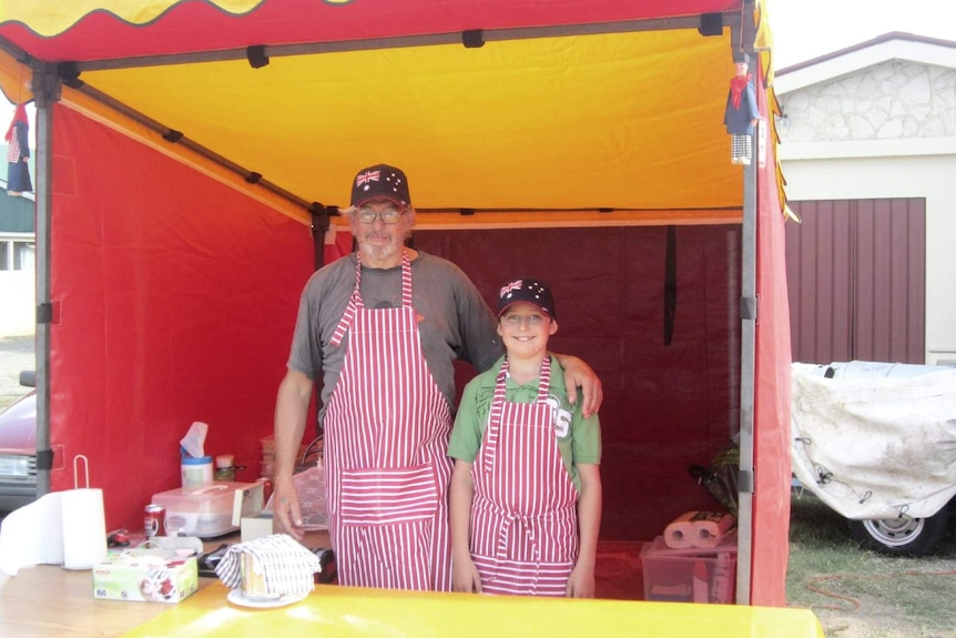 An older man and young boy wearing red and white striped aprons and Australia caps stand next to each other smiling.