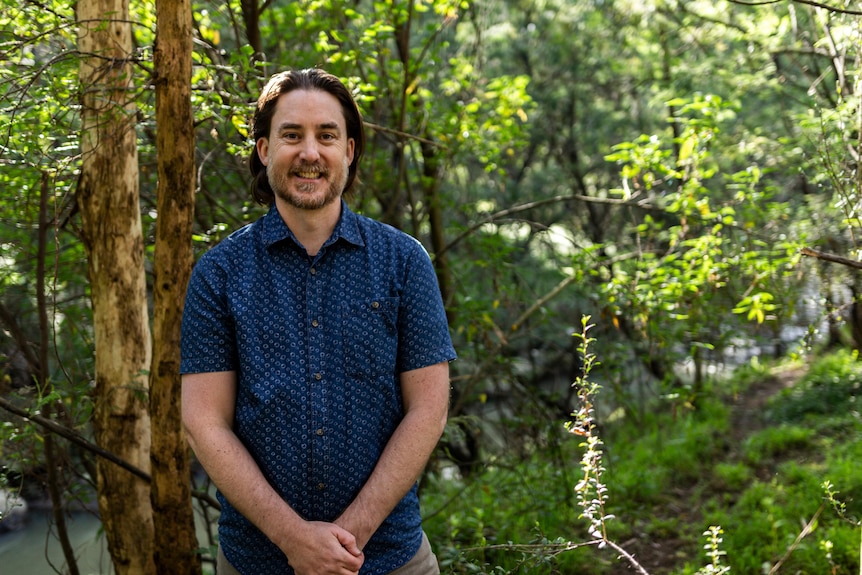 A man stands with his arms folded in front of a forest. 