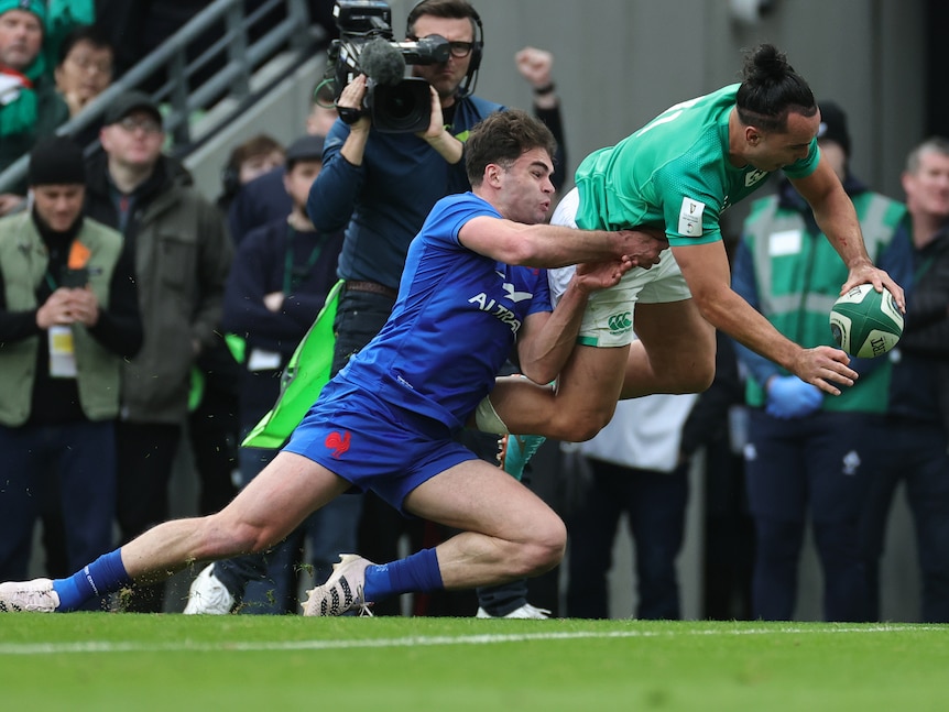 A French rugby union defender grimaces as he tries to tackle an airborne Irish player holding the ball to plant for a try.