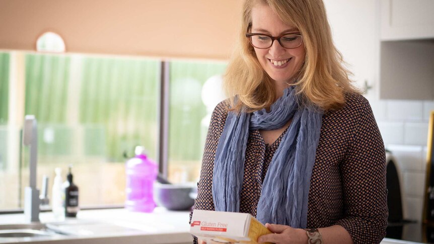 A woman looks at packaging emblazoned with ‘gluten free’ on the box, standing in a kitchen.
