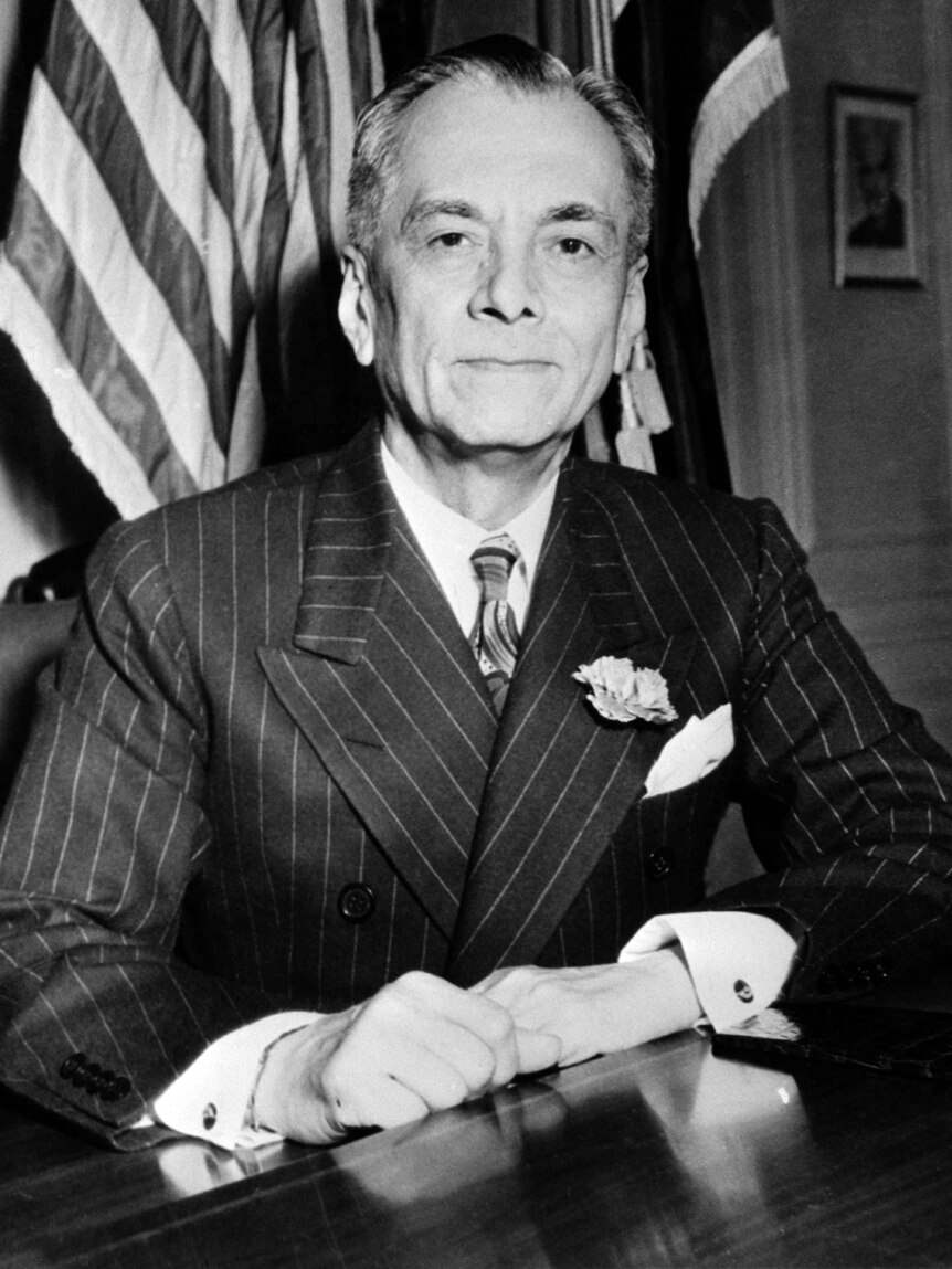 A distinguished-looking man in a suit sits at a desk in front of flags.