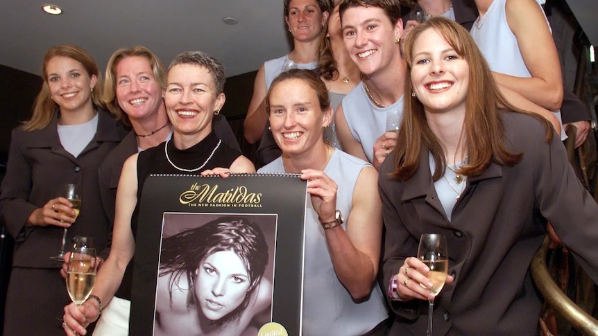Women wearing grey suits pose for a photo while holding champagnes and a calendar