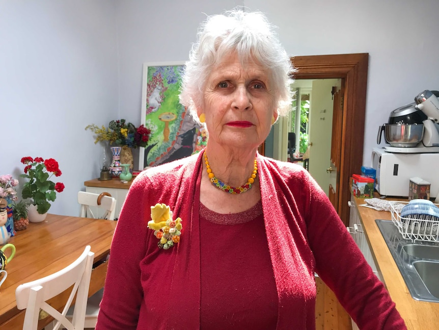 An elderly lady with white hair lit from the top with natural light wearing a red cardigan with a yellow brooch in her ktichen