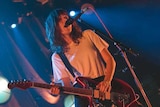 A woman with brown hair and wearing a white t-shirt stands on a stage playing guitar and singing into a microphone