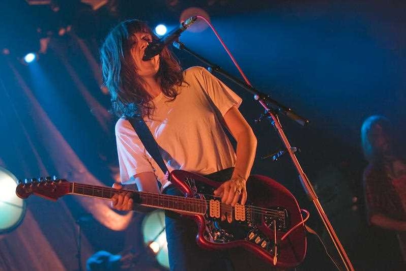 A woman with brown hair and wearing a white t-shirt stands on a stage playing guitar and singing into a microphone