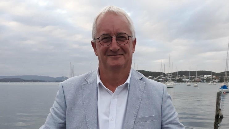 A white-haired man with glasses wearing a pale grey suit jacket stands with his back to a harbour.