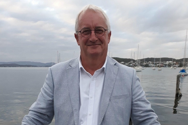 A white-haired man with glasses wearing a pale grey suit jacket stands with his back to a harbour.