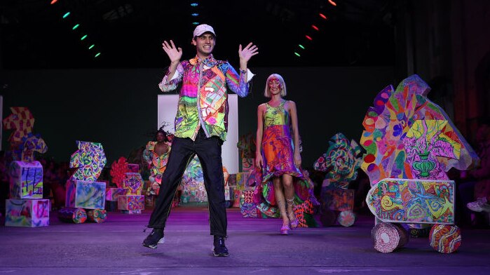 Jordan Gogos waves while wearing a brightly coloured shirt and black pants, surrounded by cardboard sculptures in a warehouse.