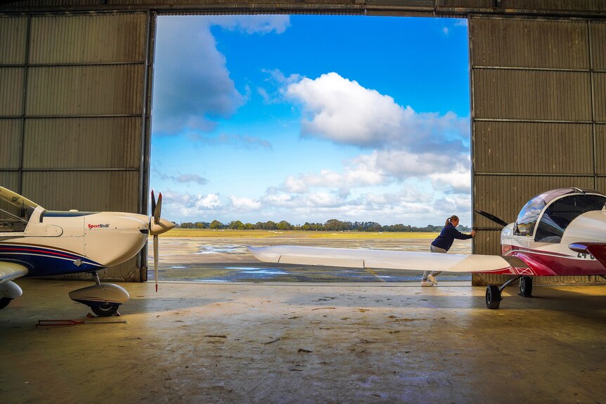 A teenage girl pushes open a hangar door to reveal a stretch of tarmac on a sunny day.
